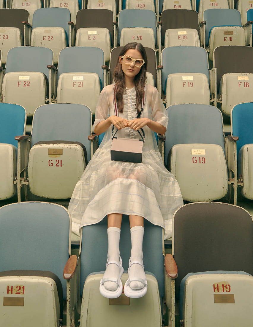 A model in velcro strap sandals, a top handle box bag and printed geometric frame sunglasses – sitting down.