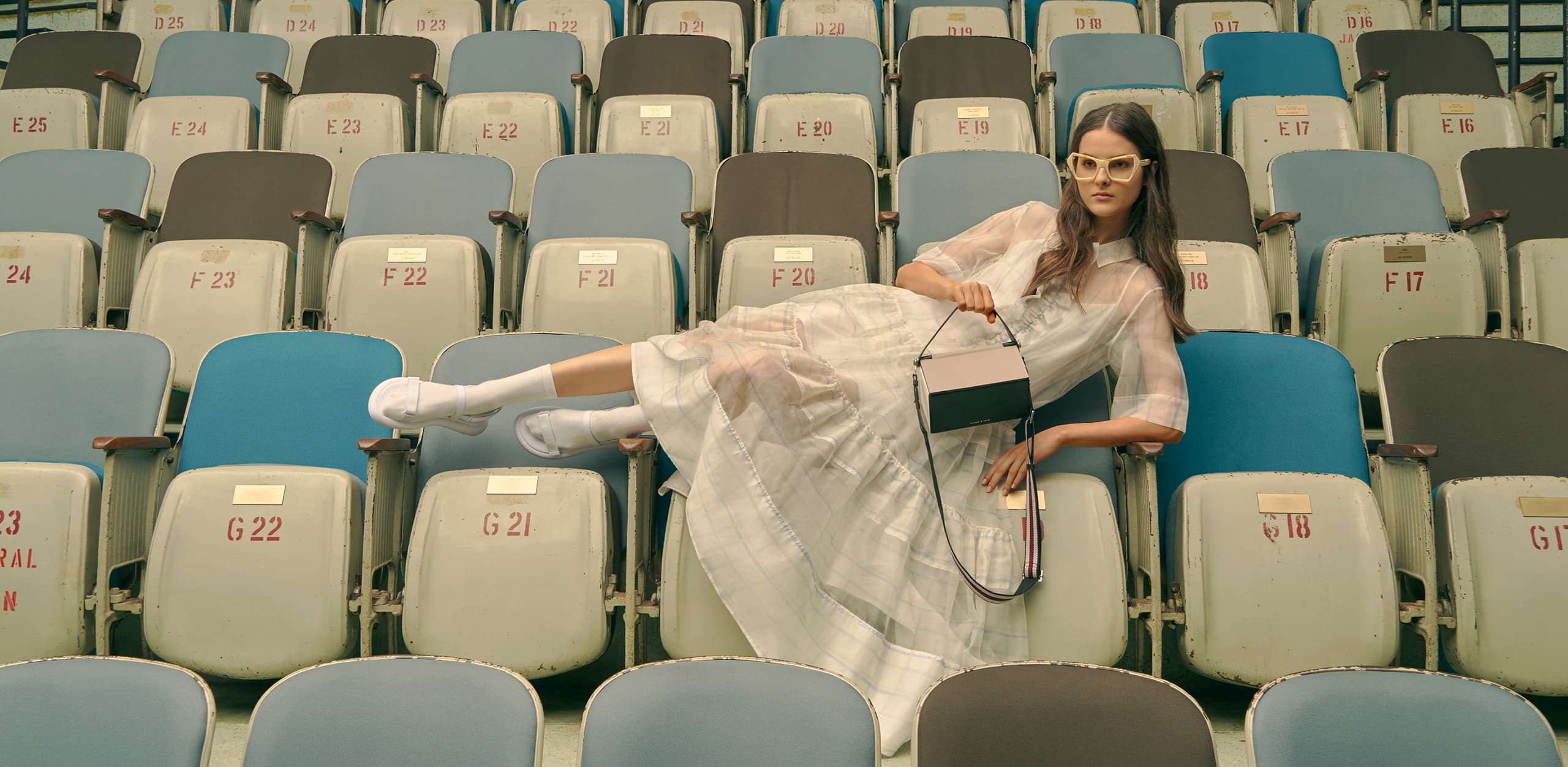 A model in velcro strap sandals, a top handle box bag and printed geometric frame sunglasses.